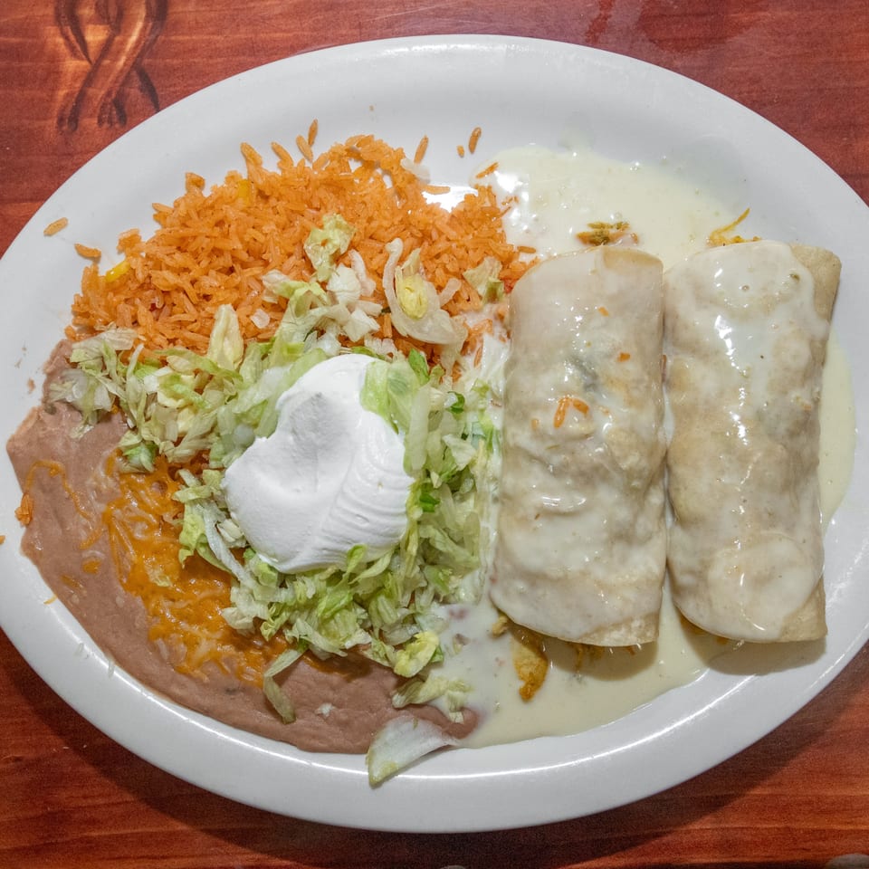 Two enchiladas covered in white sauce, with rice, refried beans, lettuce, and sour cream on a white plate.