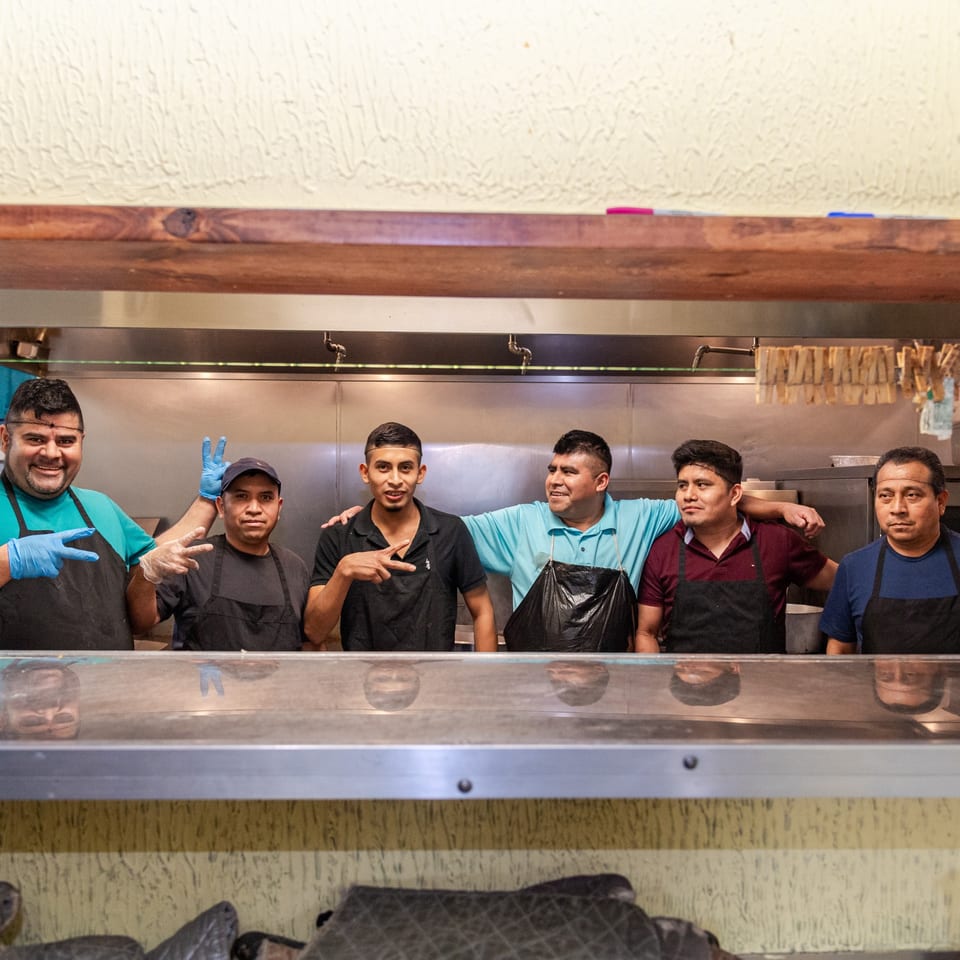 Six smiling Mi Mexico Restaurant kitchen staff members in aprons and gloves pose behind a counter.
