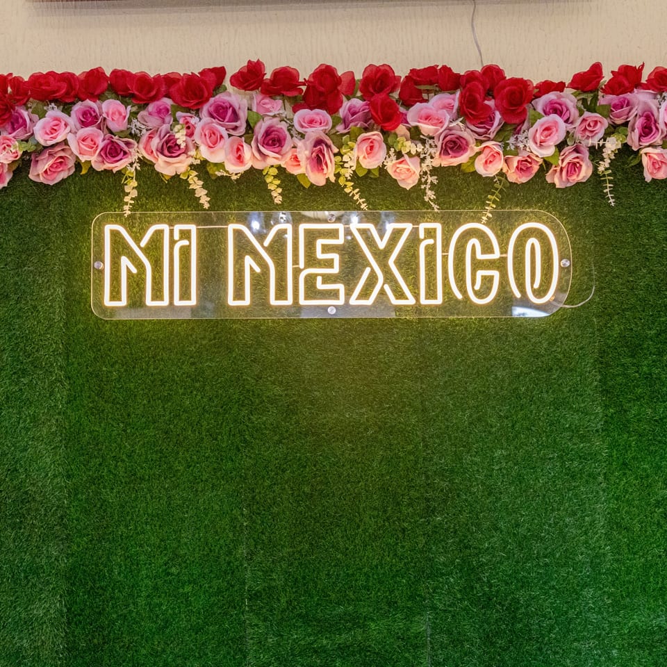 Neon "Mi Mexico" sign on a green wall, adorned with a garland of pink and red roses.