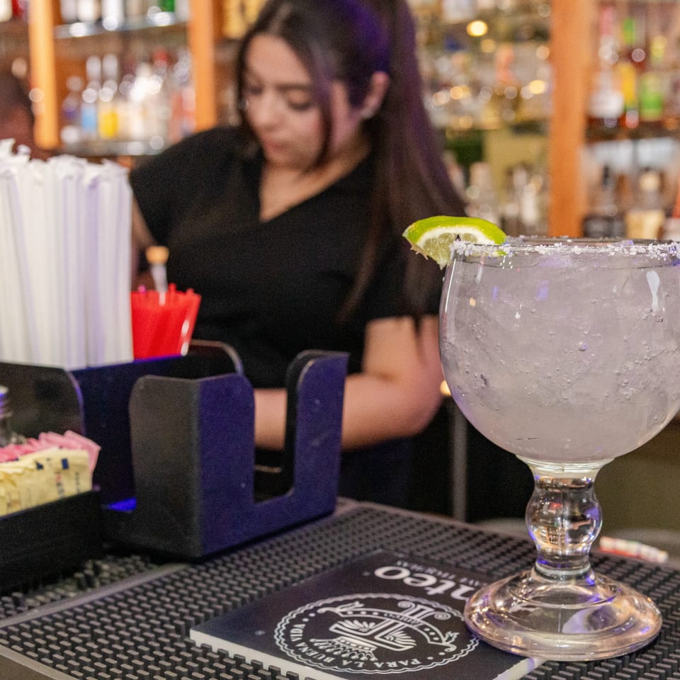 A bartender prepares drinks behind a bar, with a large, salted margarita in the foreground.