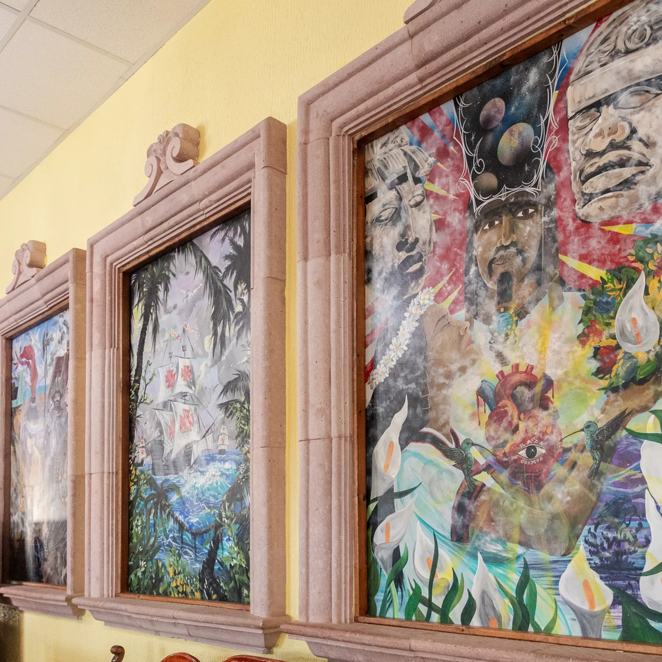 Three framed paintings on a yellow wall, depicting Mexican cultural and historical scenes.